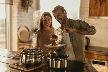 Man and woman happily serving homemade pasta at a dining table in a sunlit kitchen