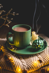 Steaming mug of hot chocolate resting on a green saucer beside an extinguished candle and ornament,...