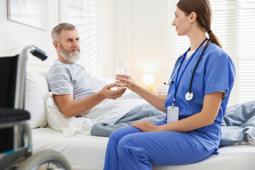 Caregiver giving water to senior man on bed indoors. Home health care service