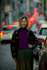 A smiling woman in a skirt and a warm checkered jacket stands on a busy evening street, surrounded...