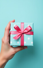 My hand touches a charming pastel blue gift box adorned with polka dots and a sweet pink ribbon bow on a matching canvas background._00001_