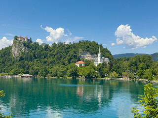 Fototapeta premium Lake Bled, Slovenia - June 28, 2024: St. Martin Church with gray-white mosaic roof surrounded by green foliage under blue cloudscape. White cliffs in back, on top the castle