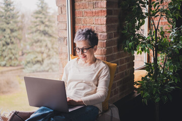 Charming senior woman with short grey hair enjoying leisure time at home, using laptop near window in cozy daylight apartment