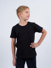 Pleased blonde haired boy points at blank black t shirt shows place for your advertisement focused down isolated over white background. People clothing and promotion concept.