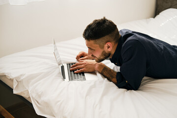 Obraz premium A young Caucasian bearded man is lying on a bed with a laptop. Working online from home concept. The guy is a freelancer at home interior.
