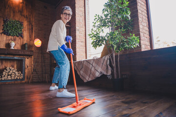 Charming mature woman joyfully cleaning wooden floor at home in daylight, wearing casual pullover...