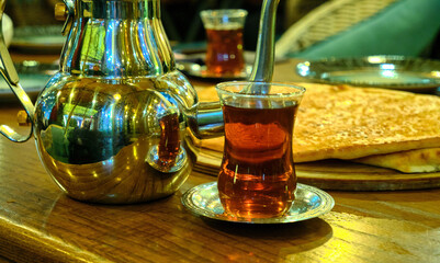 A traditional Turkish café scene with tea in a curved glass cup, a metal saucer, a shiny teapot, and bread in the background.