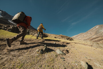 Tourists go uphill with backpacks, in hiking clothes and shoes. The view from the side goes to the mountains. Travelers with backpacks. Hiking boots.