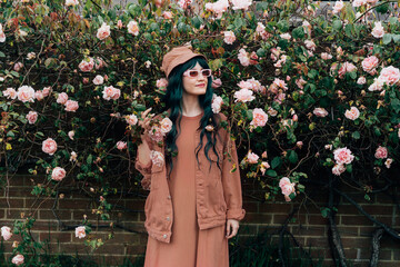 Portrait of young hipster woman in mocha color dress, jacket, bucket hat and sunglasses on blooming roses bush background. Urban street fashion. Gen Z. Trendy fashion outfit. Color of the 2025 year.