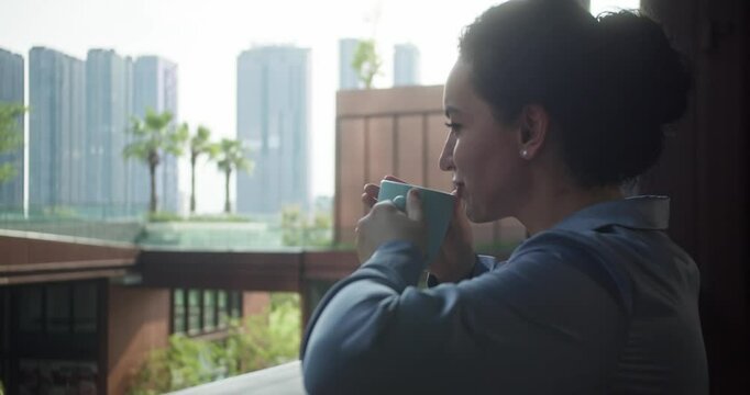 Young Indian woman standing on her terrace against the backdrop of skyscrapers and multi-story buildings, enjoying drinking coffee, watching views of sunrise landscape and scenery.
