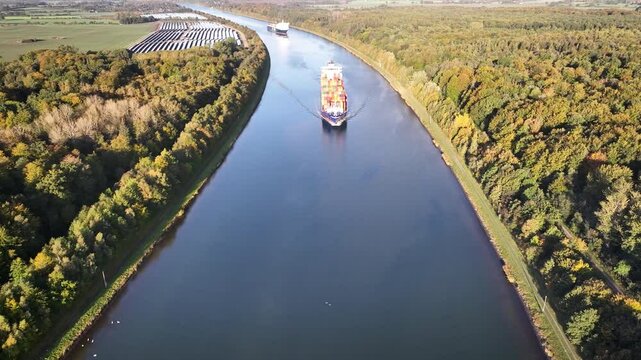 Container ship and car ferry navigate the Kiel Canal alongside vast solar panel fields and wind turbines, set against a backdrop of forest.