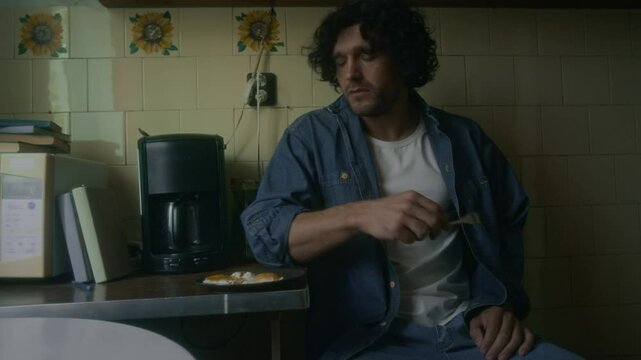 Medium shot of depressed Caucasian man taking seat at table to have breakfast in kitchen, eating fried eggs in morning