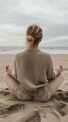 Fototapeta premium A woman with her hair tied back sits cross legged on the sandy beach, wearing a cozy sweater