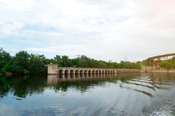 Obraz premium A serene view of an old stone bridge with multiple arches spanning a calm river, surrounded by lush green trees and a clear sky, perfect for nature and architecture enthusiasts