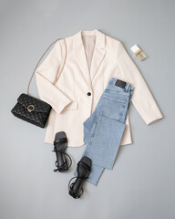 Beige jacket, blue jeans, black cross body bag with chain, black heeled sandals on grey background....