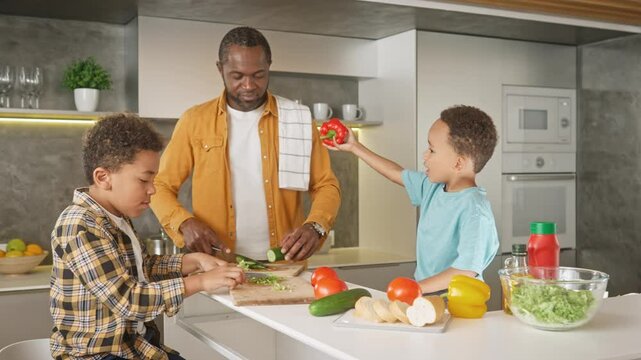 Happy African American father chopping vegetables with his son, while another kid serving fresh peper for them. Enjoying preparing healthy salad. Cooking together in modern kitchen.