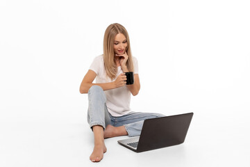 Fototapeta premium A young woman enjoys a warm drink while sitting on the floor with her laptop in a bright, minimalist room during the afternoon