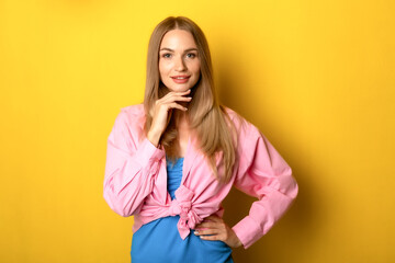 Young woman posing confidently against a vibrant yellow backdrop, dressed in a blue dress and a pink cropped jacket