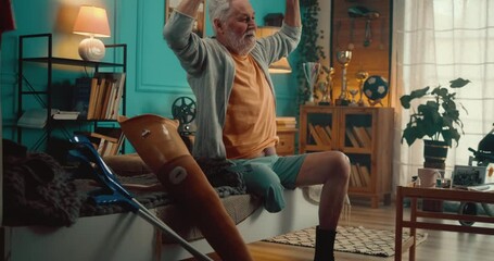 An elderly senior man with a disability, a former football player with a Prosthetic Leg sits on the bed in the morning and stretches in his room with a soccer ball and trophies in the background - Powered by Adobe
