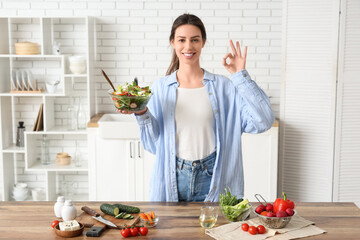 Beautiful young woman with glass bowl of fresh vegetable salad showing ok gesture in kitchen