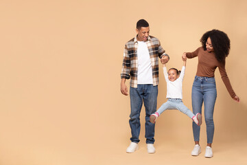 Funny black parents playing with daughter, holding hands and raising girl up, family posing over beige studio background, full length shot, copy space