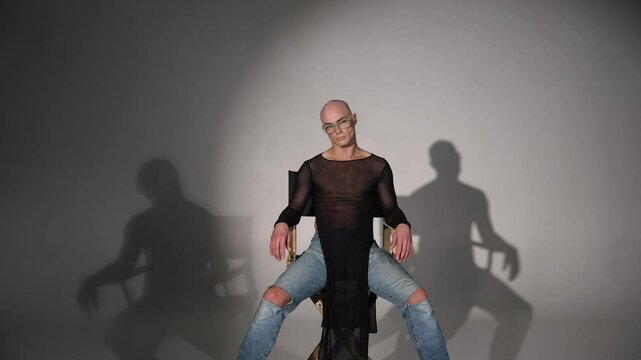A guy - a dancer sits on a chair and starts dancing. A guy dances a modern dance, on a chair in a studio. An outrageous man in glasses sits on a chair and dances.