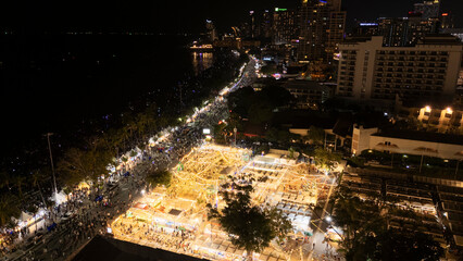 Aerial view of a vibrant nighttime festival market illuminated by warm string lights, set along a lively beachfront and surrounded by a bustling cityscape.