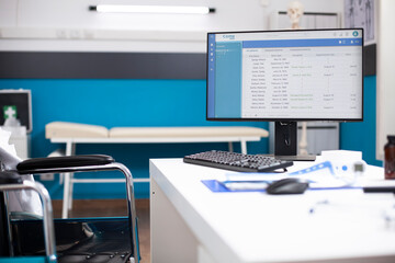 Empty wheelchair user physician desk with computer screen displaying patient health records for upcoming appointments. Clinical setting with examination tools and organized medical documents.