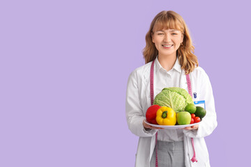 Young happy female nutritionist with fresh vegetables and measuring tape on purple background