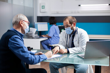 Fototapeta premium General practitioner aids elderly caucasian man in filling out medical information on clipboard. Young doctor and male pensioner, both wearing masks, discussing health assessment in clinic office.