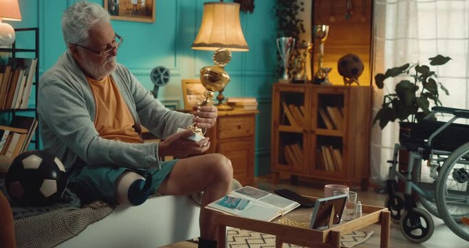 Former 70s male soccer player with an amputated leg holding and kissing atrophy, showing sign of victory with his fists, admiring and recalling his past and youth looking at photos and his won tro