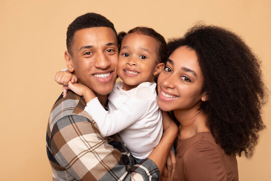 Happy African American parents hugging their pretty little daughter and smiling together to camera , posing on beige background, studio shot