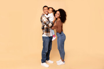 Excited young African American family of three posing on beige background, father holding little daughter on hands, family expressing happiness and love