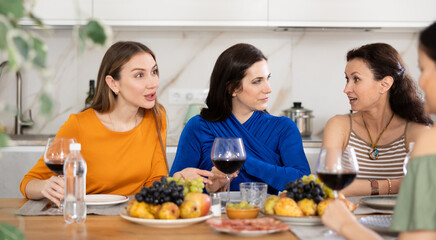 Cheerful female friends enjoying home gathering with wine and light snacks at kitchen table, engaging in lively conversations..