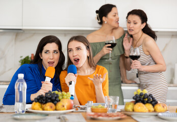 Female participants of friendly feast sing their favorite songs in karaoke and have fun. Two friends whisper and share secrets, talking with glasses in their hands in background