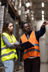 Warehouse workers in safety vests engage in discussion, reviewing logistics strategy in an industrial setting. The man points while holding a clipboard, 