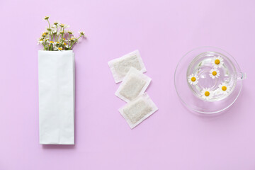 Chamomile flowers, tea bags and cup with water on lilac background. Top view