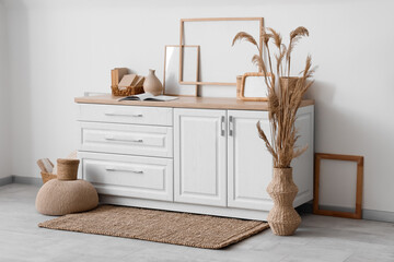 Stylish commode with different home decor and vase of dry reeds near white wall in room