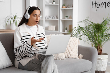 Shocked young woman in headphones with laptop video chatting on sofa at home