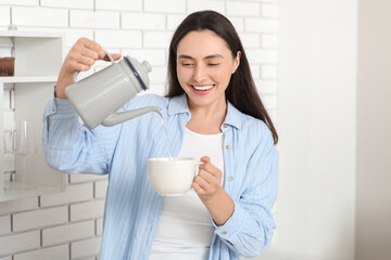 Beautiful young happy woman pouring hot water from kettle into cup in kitchen