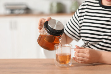 Beautiful young woman pouring hot tea from transparent kettle into cup in kitchen, closeup