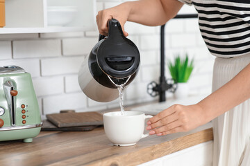 Beautiful young woman pouring hot water from modern electric kettle into cup in kitchen, closeup