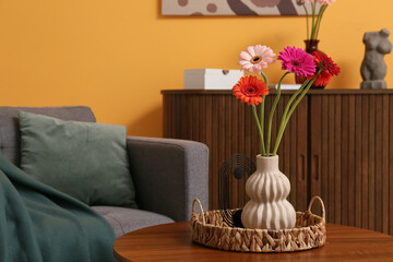 Armchair with plaid, cushion and gerbera flowers in vase on coffee table in living room. Closeup