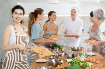 Young participiant of cooking master class holding cutting board with raw chicken breast in her hands standing around other female members