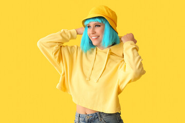 Beautiful young woman in blue wig and stylish panama on yellow background