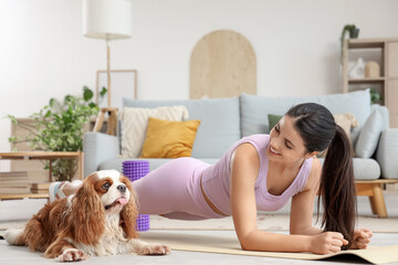 Sporty young woman exercising with cute cavalier King Charles spaniel on yoga mat at home