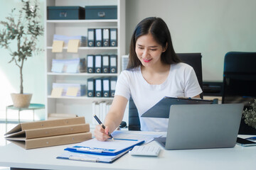 A young Asian woman works at her desk in an office room during the day. Surrounded by financial charts, files, and devices, she consults on credit reports, calculations, and credit risk assessments.