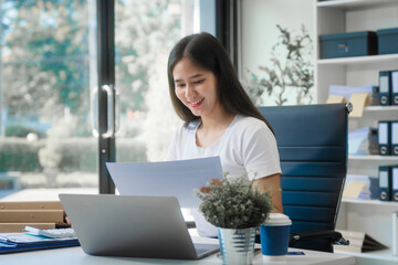 A young Asian woman works at her desk in an office room during the day. Surrounded by financial...