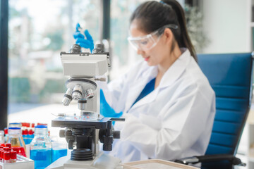 A young Asian woman works in a laboratory, conducting chemical and blood tests. Wearing goggles,...