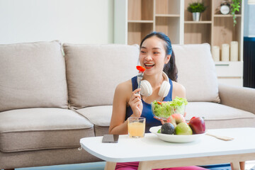 A young Asian housewife enjoys a healthy lifestyle at home, balancing fitness routines with relaxation. She sits on a modern sofa, eating a fresh salad and fruit while embracing healthy habits.